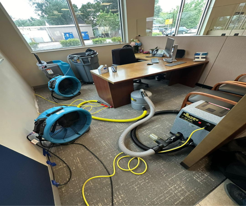 Interior of an office with an L-shaped desk and two chairs in front of it with industrial fans and other water restoration equipment on the floor