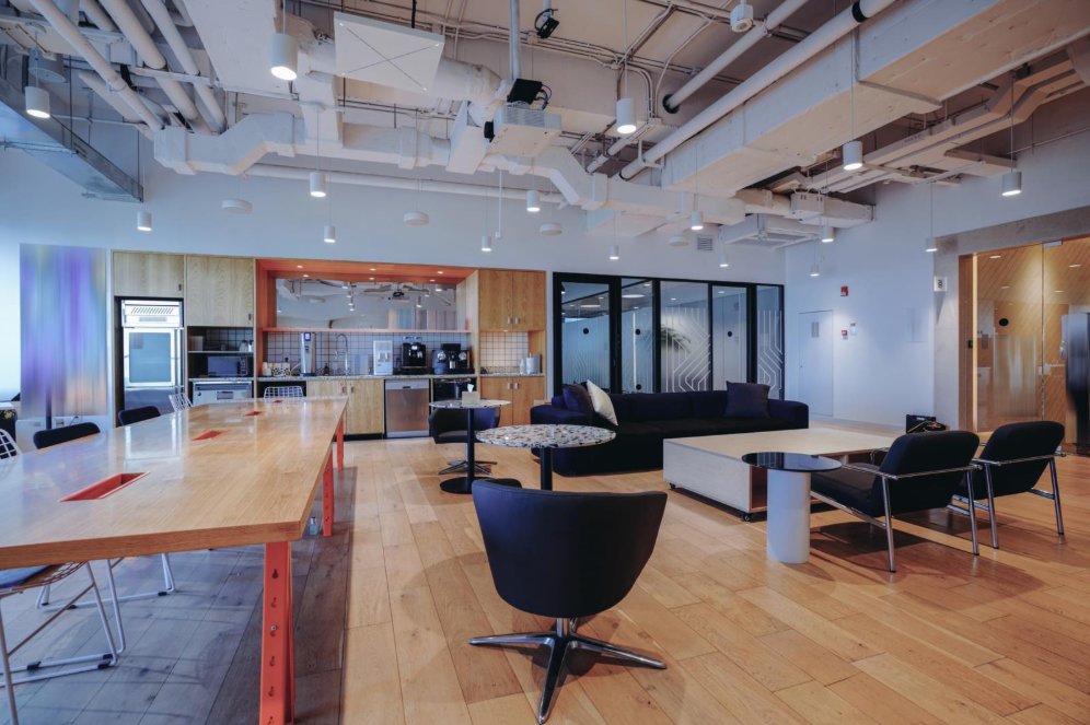 A modern office lounge area with a long table, a couch and chairs around a coffee table, and swivel chairs next to cafe tables with a coffee station and appliances in the background.